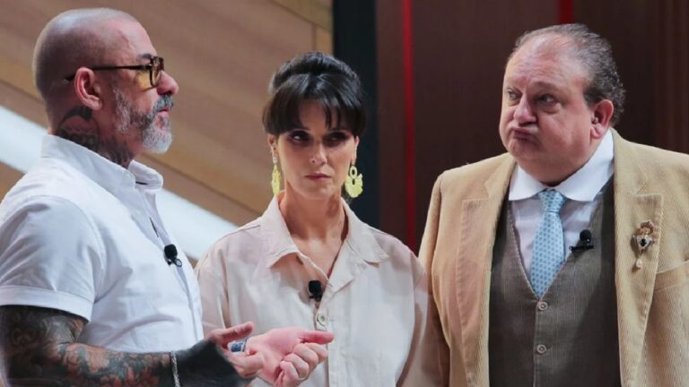 Três participantes conversam em estúdio de programa de TV, usando microfones de lapela; um homem tatuado fala enquanto mulher ao centro observa com expressão séria e outro homem de terno reage com expressão exagerada.