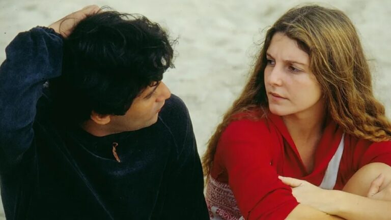 Homem e mulher estão sentados lado a lado ao ar livre, sobre a areia, em possível cenário de praia. Ele, com cabelo escuro e blusa preta, apoia a mão na cabeça enquanto olha para ela. Ela, com cabelos longos castanhos e blusa vermelha, mantém os braços cruzados sobre os joelhos e retribui o olhar com expressão séria, sugerindo uma conversa tensa ou reflexiva.