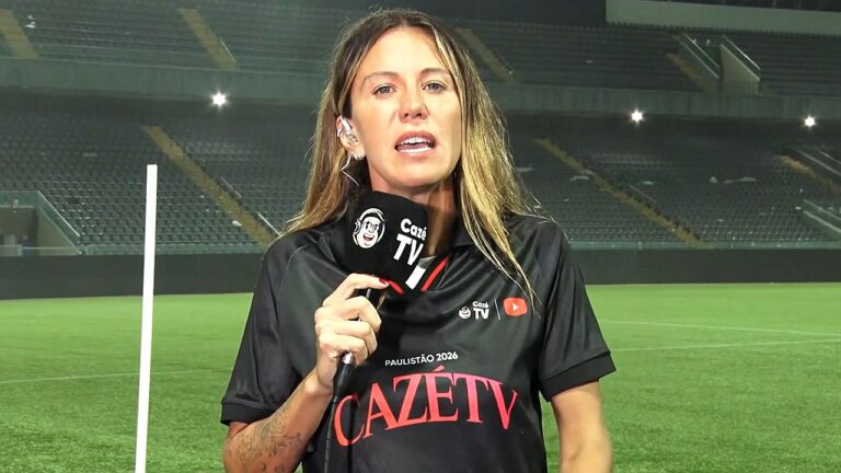 Repórter esportiva aparece em campo de futebol vazio, segurando microfone com a marca “Cazé TV”. Ela veste camisa preta com a inscrição “Paulistão 2026” e está posicionada diante das arquibancadas iluminadas do estádio, indicando cobertura jornalística pós-jogo ou pré-partida.