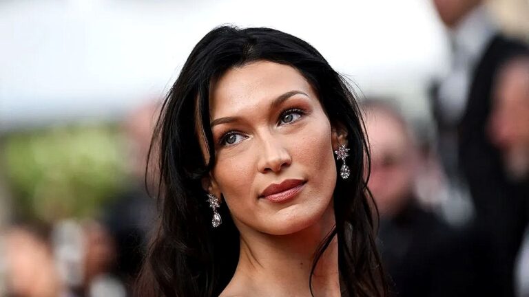 Mulher com cabelo longo e escuro posa em evento de tapete vermelho, usando brincos brilhantes e maquiagem suave. Ela olha levemente para o lado, com expressão serena, enquanto o fundo desfocado sugere fotógrafos e convidados ao redor.