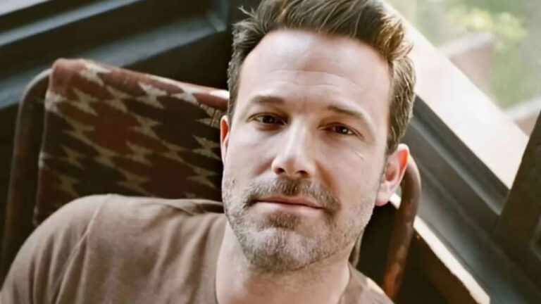 Ator Ben Affleck de barba rala e camiseta marrom olha seriamente para a câmera sentado em cadeira com encosto de estampa étnica perto de uma janela