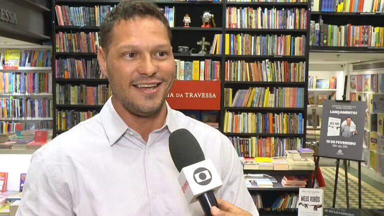 Homem concede entrevista em uma livraria, com estantes repletas de livros ao fundo. Ele veste camisa social clara e sorri enquanto fala ao microfone com logotipo de emissora de TV. Ao lado, há um cartaz de lançamento de livro exposto em cavalete, indicando evento literário no local.