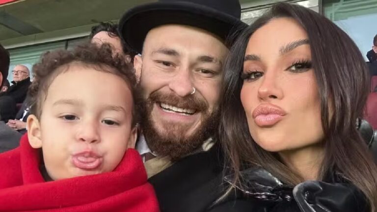 Criança enrolada em manta vermelha posa ao lado de homem sorridente e mulher fazendo biquinho em arquibancada de estádio