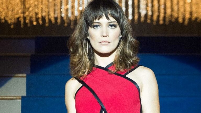 Mulher de cabelos castanhos na altura dos ombros e franja reta posa com expressão séria em ambiente interno iluminado por luzes douradas ao fundo. Ela veste um vestido vermelho sem mangas com detalhes em preto cruzando a parte superior. A imagem sugere evento social ou desfile, com fundo desfocado e iluminação sofisticada.