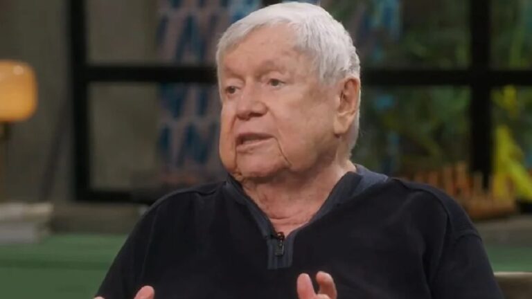 Homem idoso de cabelo branco e blusa escura fala durante entrevista em estúdio de TV com cenário desfocado ao fundo