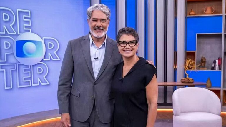 Dois apresentadores posam sorrindo em estúdio de televisão. O homem, de terno cinza e camisa clara, está com o braço ao redor da mulher, que usa vestido preto e óculos de armação escura. Ao fundo, há painel azul com parte do logotipo do programa e um cenário com poltrona, estante e objetos decorativos, caracterizando ambiente de talk show