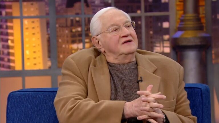 Homem idoso de óculos e casaco bege conversa sentado em sofá azul durante entrevista em programa de TV