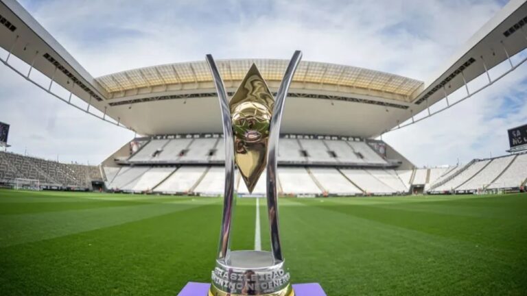Troféu do Campeonato Paulista posicionado no centro do gramado de um estádio de futebol vazio, com arquibancadas brancas e cobertura ampla ao fundo.