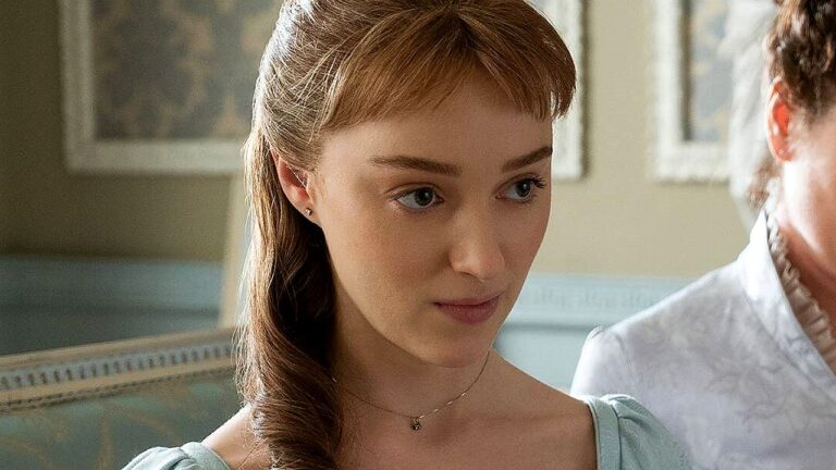 Mulher jovem com cabelos castanhos presos em penteado semi-preso aparece em ambiente interno com decoração clássica. Ela veste vestido em tom claro e colar delicado, olhando levemente para baixo com expressão atenta e contida. Ao fundo, há paredes com molduras e outra pessoa parcialmente visível, sugerindo cena de diálogo em contexto histórico ou dramático.