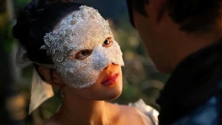 Mulher usa máscara prateada e texturizada que cobre a região dos olhos e parte do rosto, olhando fixamente para um homem à sua frente. A cena, em close, tem iluminação quente e fundo desfocado, sugerindo momento íntimo ou misterioso em ambiente interno.