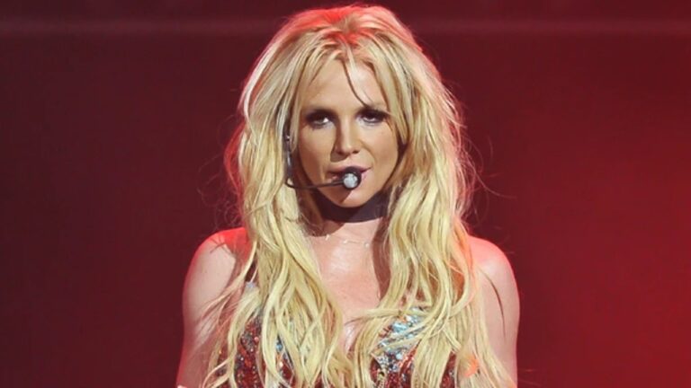 Mulher de cabelos loiros longos se apresenta no palco com microfone de cabeça e iluminação vermelha ao fundo.