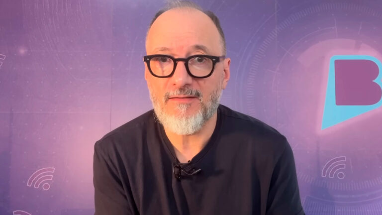 Britto Jr de óculos pretos e barba grisalha fala diante de fundo roxo com logotipo estilizado ao fundo em estúdio de gravação