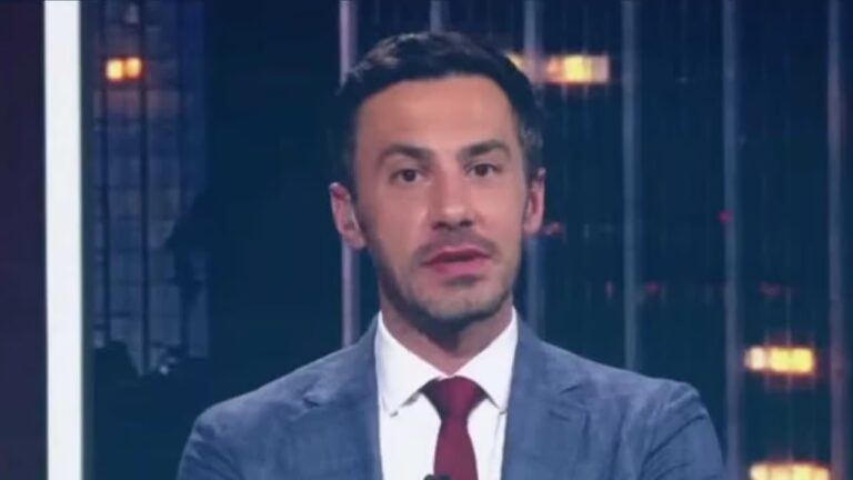 Homem aparece em estúdio de TV com expressão séria, olhando diretamente para a câmera. Ele veste terno azul, camisa branca e gravata vermelha, com microfone preso à lapela. Ao fundo, há iluminação baixa e painéis com luzes e estruturas que sugerem cenário de telejornal ou programa de análise.