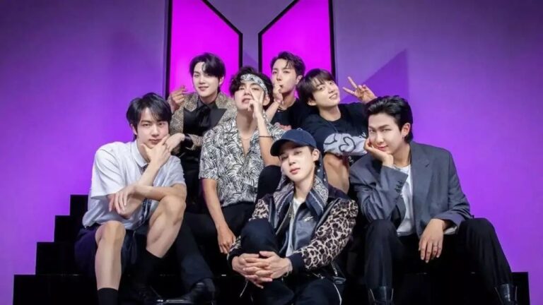 Sete integrantes de grupo musical posam sentados em degraus com fundo roxo e logotipo estilizado em formato de M