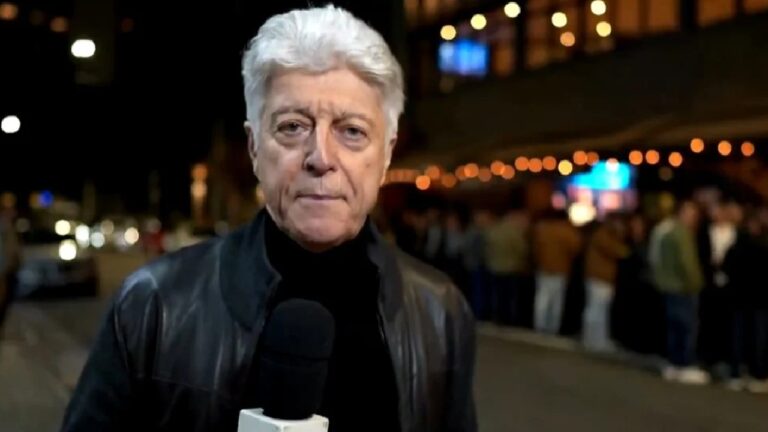 Repórter de cabelo grisalho segura microfone durante gravação noturna em rua movimentada com pessoas e luzes ao fundo