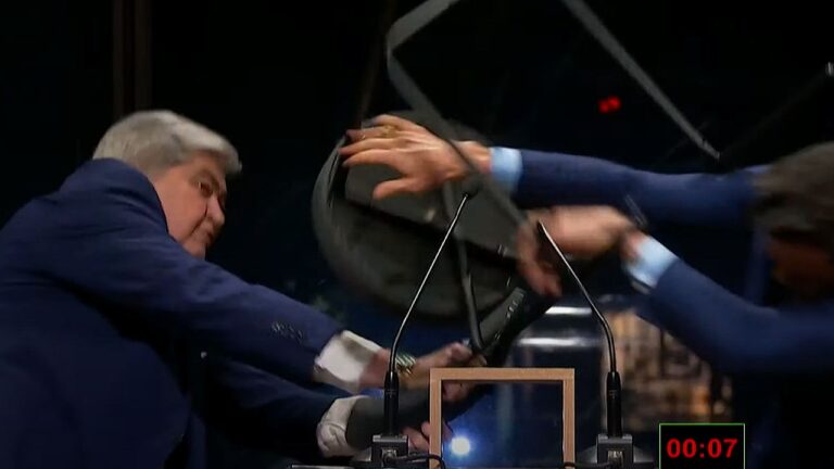 Dois homens de terno azul se inclinam sobre uma mesa em estúdio de TV enquanto disputam ou tentam segurar um objeto circular escuro. Há microfones posicionados à frente deles e um cronômetro digital aparece no canto da tela marcando sete segundos, sugerindo dinâmica ou desafio ao vivo.