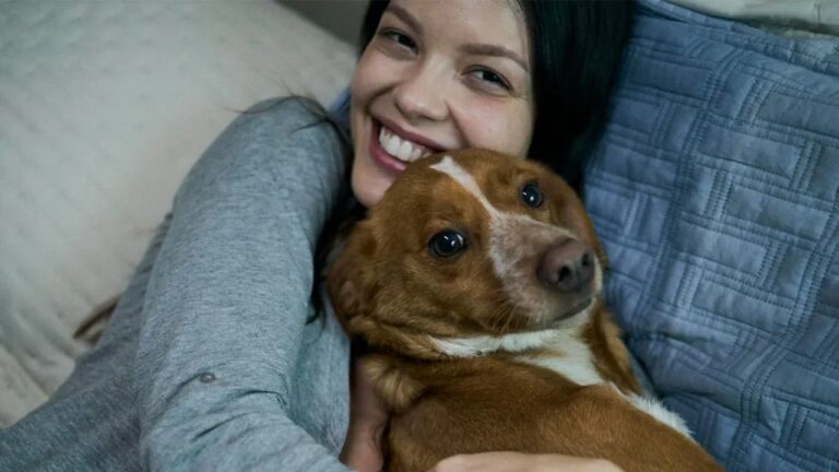 Mulher sorrindo abraça cachorro marrom deitado no sofá em cena doméstica de filme ou série dramática
