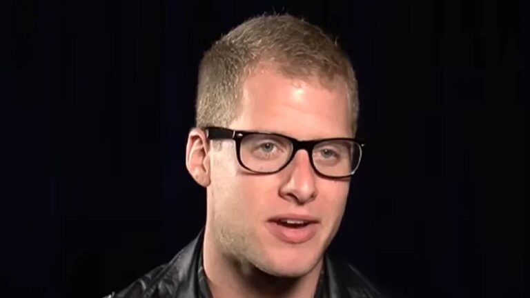 Homem jovem, de cabelo curto e claro, usa óculos de armação preta e jaqueta escura enquanto fala durante entrevista. A imagem é um close no rosto, com fundo preto, destacando sua expressão séria e foco na fala.