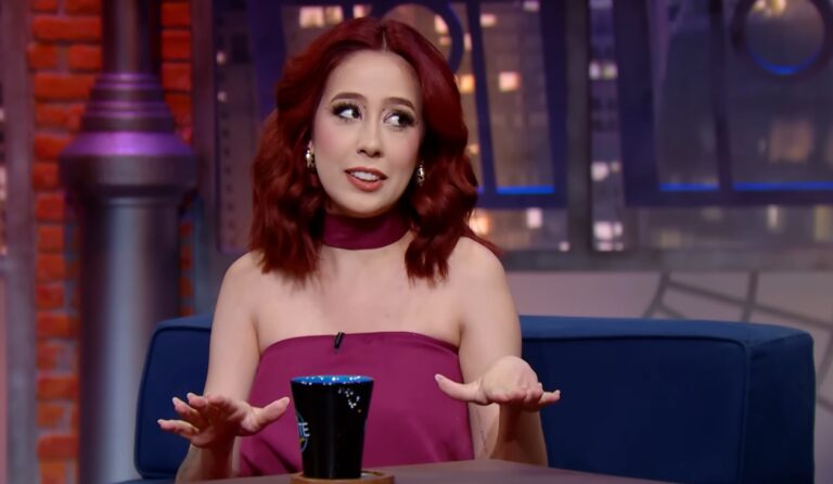 Mulher de cabelo ruivo e vestido vinho fala gesticulando durante entrevista em programa de talk show