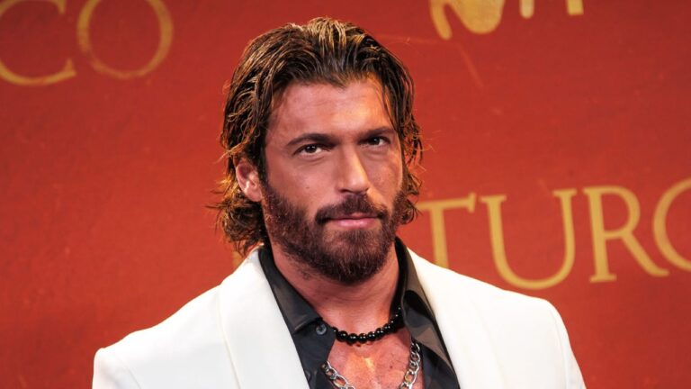 Can Yaman de paletó branco e camisa preta aberta, ostentando cabelos longos ondulados, barba cheia e colares de contas e corrente em evento