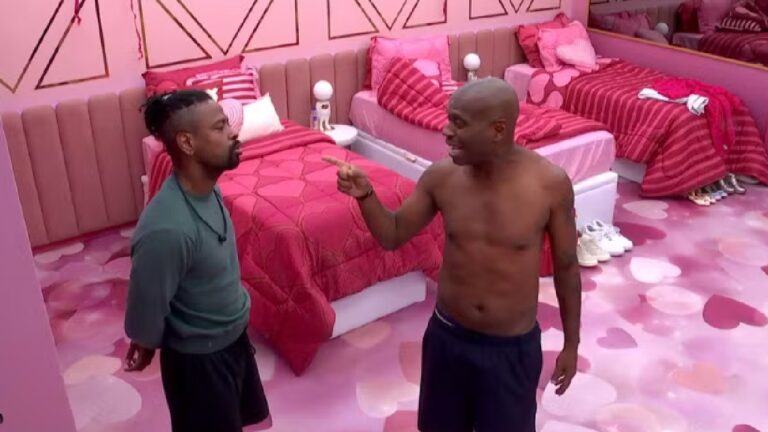 Dois homens discutem em um quarto com decoração rosa e temática de corações; um deles, sem camisa, aponta o dedo para o outro, que mantém postura séria ao lado de camas alinhadas no ambiente.