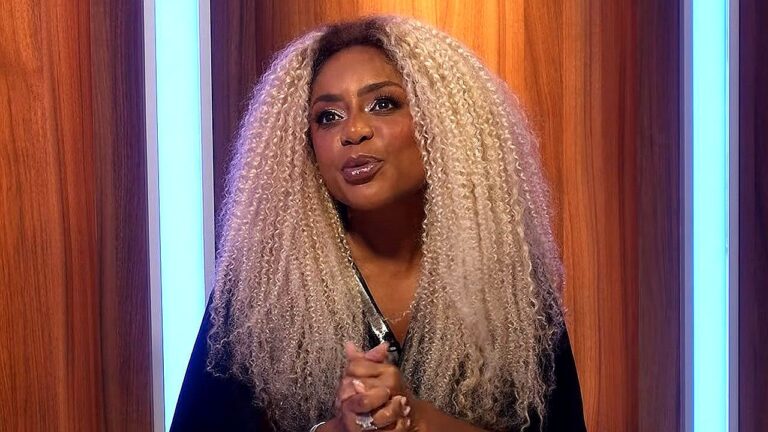 Mulher com cabelo longo, volumoso e cacheado em tom loiro claro aparece em estúdio de TV, usando roupa escura. Ela está com as mãos unidas à frente do corpo e expressão atenta, como se estivesse falando ou ouvindo alguém durante programa. O cenário ao fundo tem painéis verticais de madeira e iluminação em tons de azul, sugerindo ambiente de talk show ou atração televisiva.