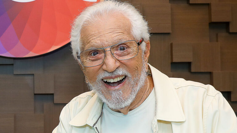 Carlos Alberto de Nóbrega sorri para a câmera, apresentador veterano de cabelos brancos e barba por fazer, veste jaqueta bege clara, fundo amadeirado