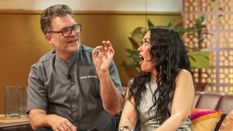 Chef de cozinha, usando uniforme cinza com o nome Carlos Bertolazzi, gesticula com os dedos em formato de 'ok' enquanto conversa com Milene Uehara uma mulher de cabelos longos e ondulados, que reage com expressão de surpresa; ambos estão sentados em sofá do estúdio de Love Taste, programa do SBT com decoração aconchegante ao fundo.