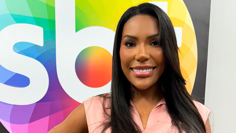 Carol Lekker sorri vestindo blusa rosa clara, posicionada à frente de um painel colorido com o logotipo do SBT em destaque ao fundo branco