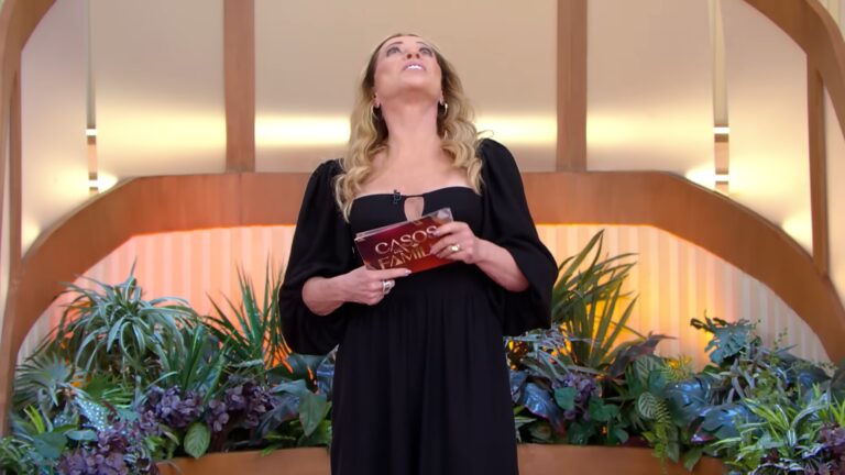 Christina Rocha, de vestido preto, segura um cartão do programa Casos de Família em estúdio decorado com plantas, olhando para cima durante a gravação, sob iluminação quente e cenário moderno