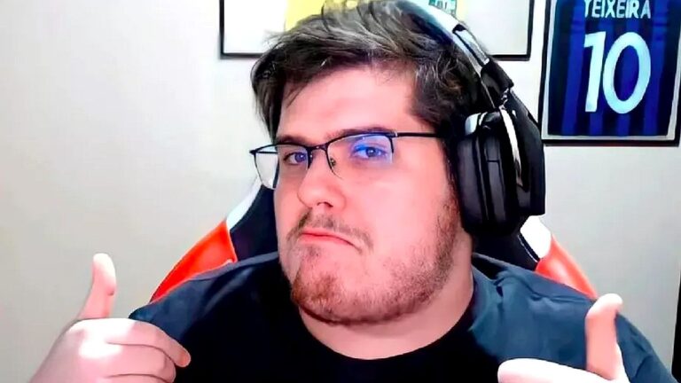 Homem usa óculos e headset enquanto faz gesto de positivo com as duas mãos diante da câmera. Ele está sentado em cadeira gamer, em ambiente interno com quadros na parede ao fundo, sugerindo gravação de vídeo ou transmissão ao vivo.