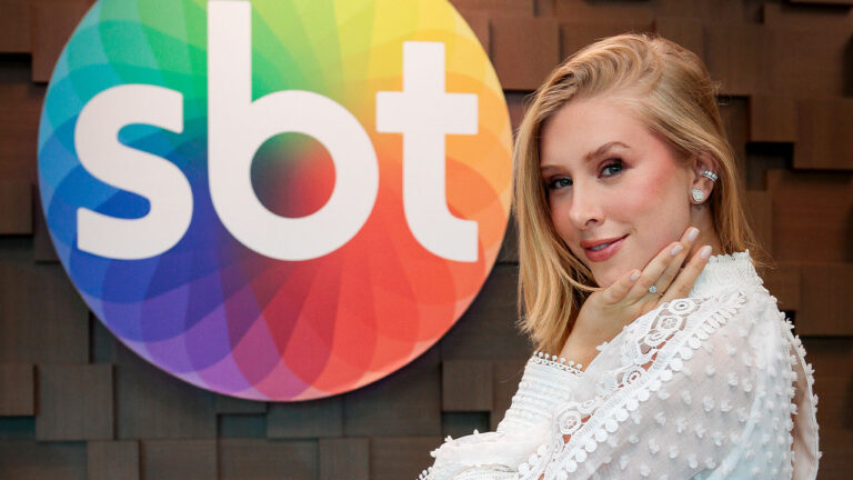 Cela Lopes de cabelos loiros e blusa branca de renda, sorridente com a mão no rosto em frente ao logotipo colorido do SBT em parede de madeira texturizada