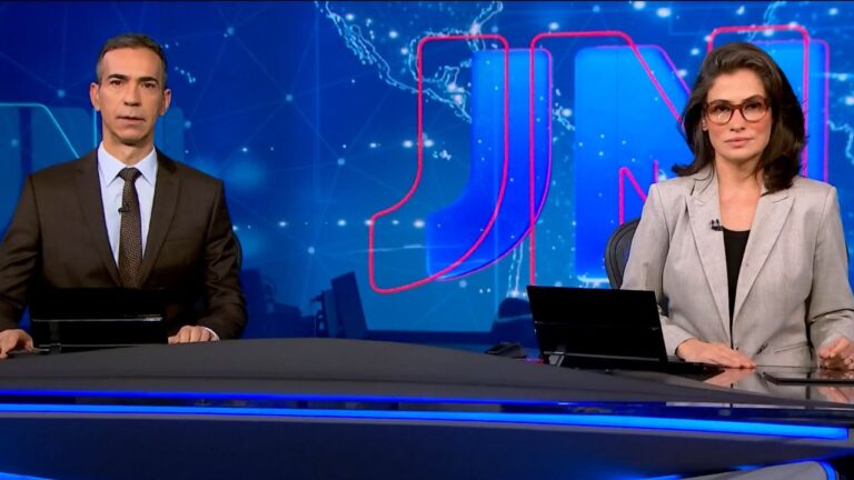 Apresentadores César Tralli e Renata Vasconcellos na bancada do Jornal Nacional, cenário com logo JN azul ao fundo, ambos usam ternos e olham fixos