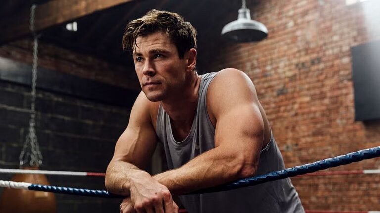 Homem musculoso apoia os braços nas cordas de um ringue de boxe, vestindo regata cinza. Ele olha para frente com expressão concentrada e determinada, em ambiente com paredes de tijolos aparentes e iluminação industrial, sugerindo treino intenso ou momento de reflexão antes de luta.