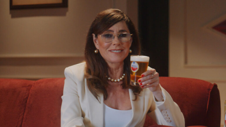 Christiane Torloni sorri de óculos e blazer branco sentada em sofá vermelho segurando copo de cerveja Amstel com colar de pérolas e brincos