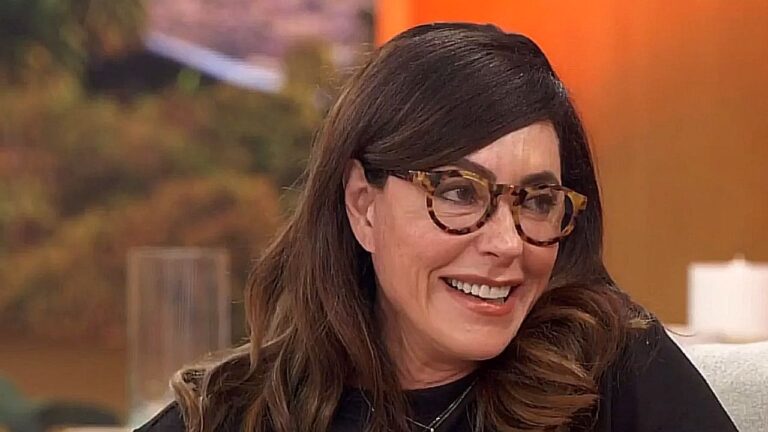 Mulher de cabelos castanhos e óculos de armação estampada sorri durante participação em programa de televisão. Ela veste blusa preta e está sentada em estúdio com fundo em tons de laranja e imagem desfocada ao fundo, em clima de entrevista descontraída.