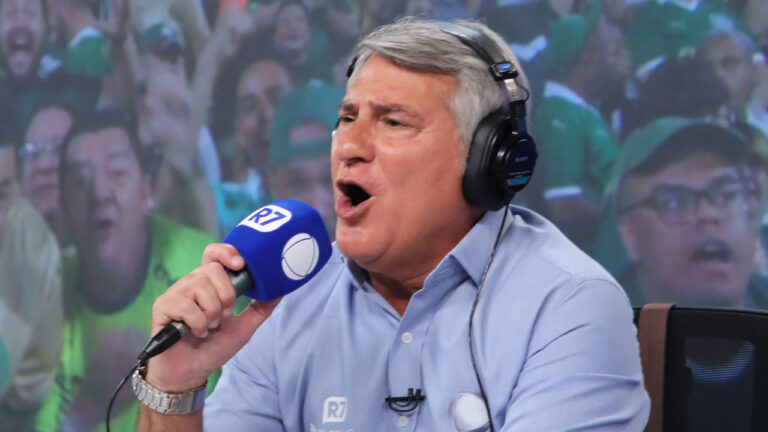 Narrador esportivo Cléber Machado narra com entusiasmo usando fones de ouvido pretos e segurando microfone azul da Record e R7, veste camisa social azul clara e fundo com torcida desfocada