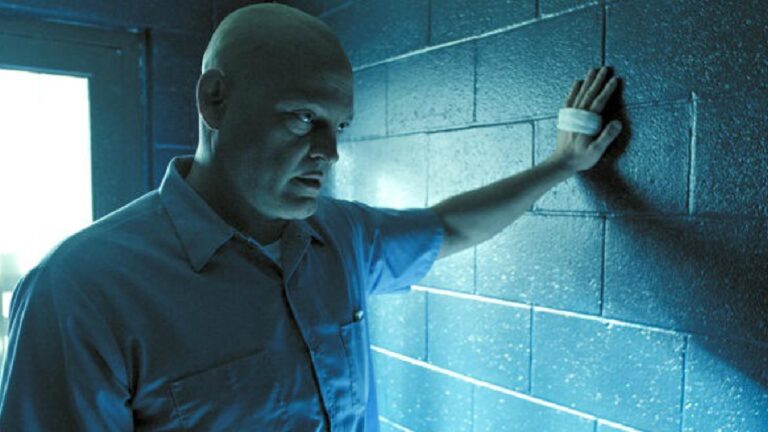 Homem careca encosta a mão na parede de blocos em ambiente azul com luz forte entrando pela porta ao fundo