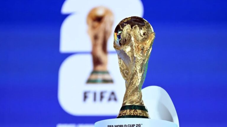 Troféu da Copa do Mundo da FIFA em destaque, com acabamento dourado e detalhes em relevo, posicionado sobre pedestal com a inscrição “FIFA World Cup 26”. Ao fundo, desfocado, aparece o logotipo da FIFA e a imagem ampliada do próprio troféu sobre fundo azul, indicando divulgação oficial do torneio.