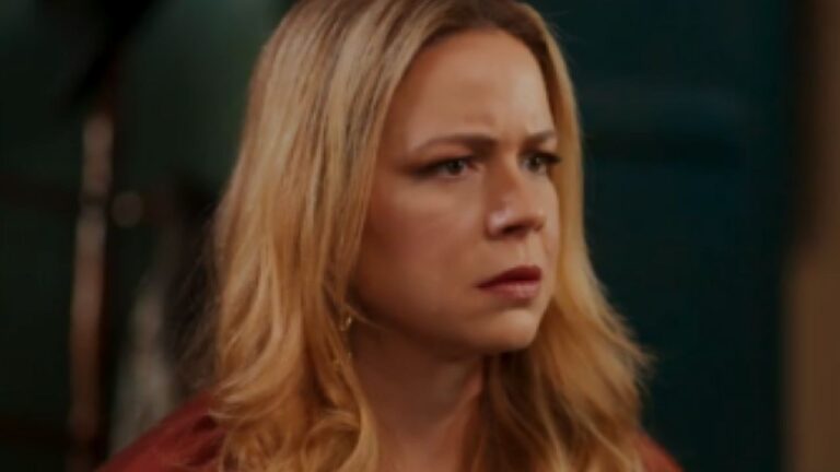 Mulher loira aparece em close, com expressão séria e olhar tenso voltado para algo fora de cena. O fundo escuro e desfocado sugere ambiente interno, indicando momento dramático em cena de novela ou série.