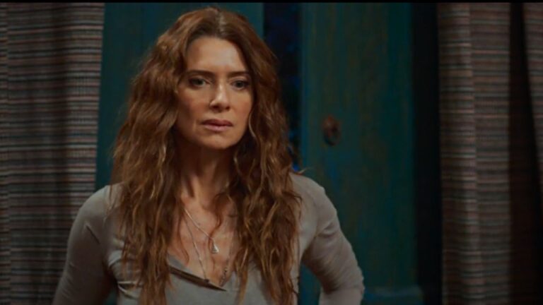 Mulher de cabelos longos e ondulados, com expressão séria e olhar atento, aparece em ambiente interno. Ela veste blusa de tom neutro e colares delicados, com a mão apoiada na cintura. Ao fundo, há uma porta de madeira pintada em verde e cortinas com listras, sugerindo uma cena de novela ou série ambientada em residência.