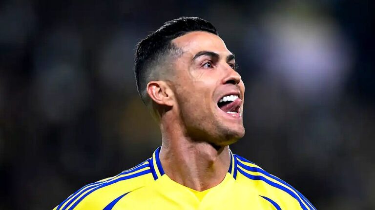 Jogador de futebol aparece em close durante partida, usando camisa amarela com detalhes azuis. Ele está com a boca aberta, em expressão intensa de comemoração ou grito, enquanto o fundo desfocado sugere estádio iluminado à noite.