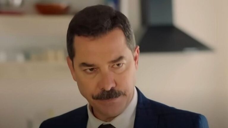 Homem de terno escuro e gravata aparece em ambiente interno, com expressão séria e olhar atento. Ele tem bigode bem aparado e está diante de cenário doméstico ao fundo, sugerindo cena de filme ou série dramática.