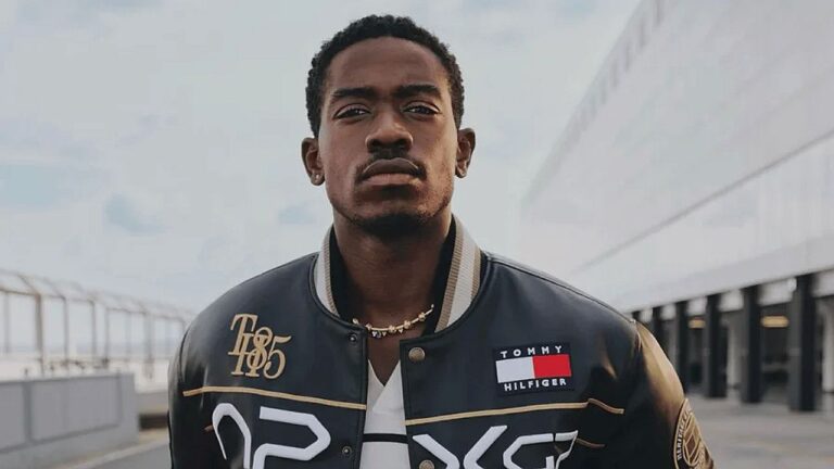 Homem posa ao ar livre com expressão séria, vestindo jaqueta estilo varsity com logotipo da Tommy Hilfiger e detalhes em dourado. Ao fundo, aparecem estruturas modernas e céu nublado, sugerindo ambiente urbano contemporâneo.