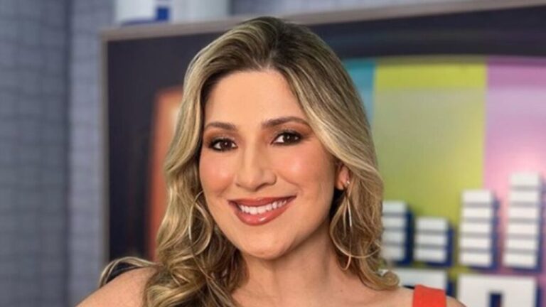 Apresentadora de cabelo loiro e vestido laranja sorri em estúdio de programa de TV com painel colorido ao fundo