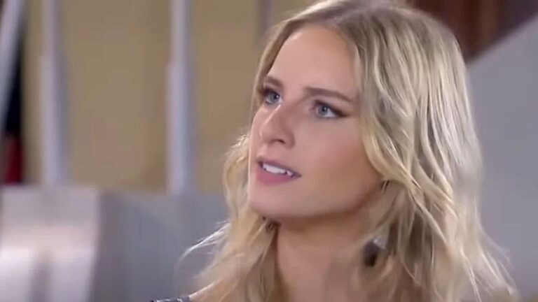 Mulher loira de cabelos ondulados aparece em close, com expressão séria e olhar atento voltado para alguém fora de quadro. A cena ocorre em ambiente interno, com fundo desfocado, sugerindo momento de diálogo intenso em produção televisiva ou cinematográfica.