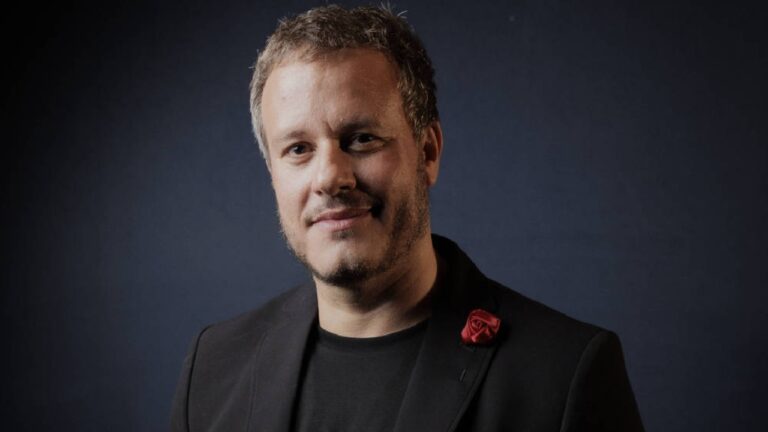 Retrato de homem com cabelo curto grisalho e barba por fazer, usando camiseta preta e blazer escuro com uma flor vermelha na lapela. Ele olha para a câmera com leve sorriso, diante de fundo azul-escuro iluminado de forma suave, em estilo de foto de estúdio.
