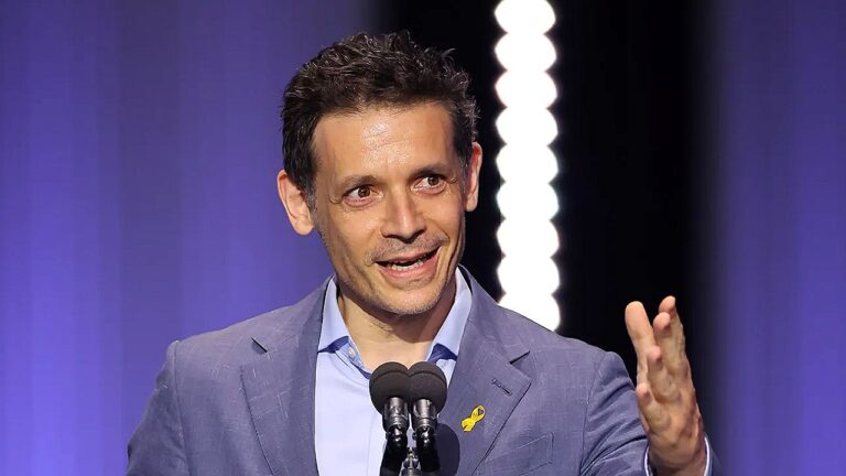 Homem fala ao microfone em evento ou premiação, vestindo blazer azul e camisa clara. Ele gesticula com a mão direita enquanto discursa, em palco com iluminação roxa ao fundo e fita amarela presa à lapela do paletó.