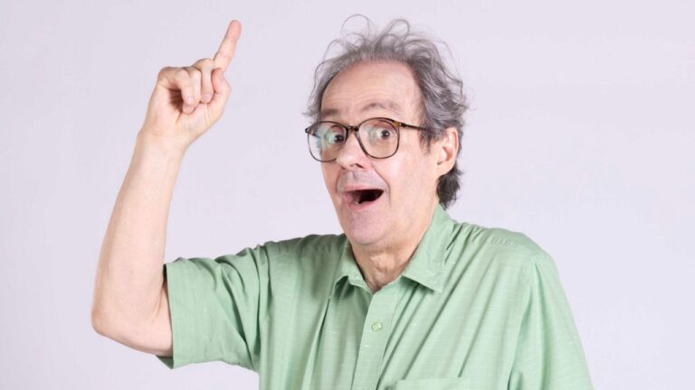 Ator Daniel Dantas usa camisa verde e óculos, posa com expressão de surpresa e aponta o dedo indicador para cima em frente a um fundo cinza claro