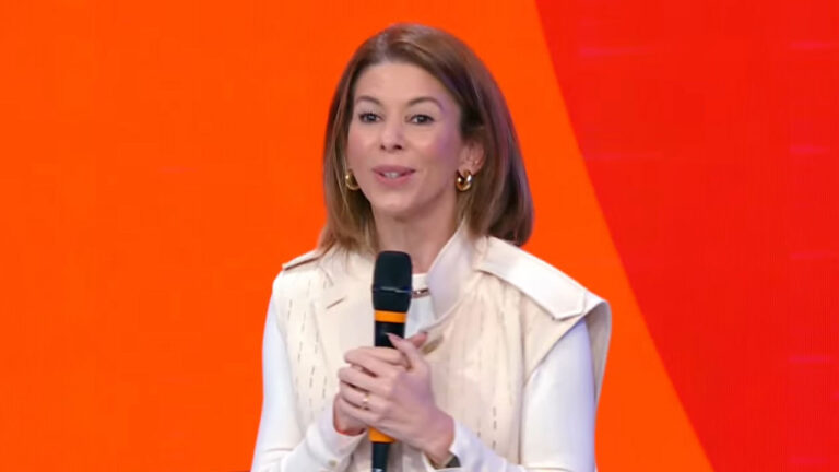 Daniela Beyruti, vice-presidente do SBT, fala ao microfone em palco, a executiva veste colete bege sobre blusa branca, usa brincos e fundo laranja vibrante